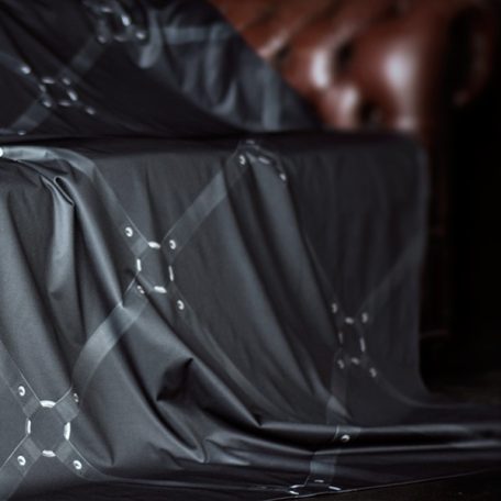 Black waterproof throw printed with a diamond pattern of black leather bondage straps with silver metal connector rings draped over part of a brown leather Chesterfield with a little of the arm showing